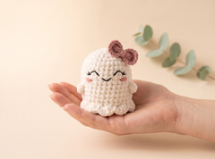 A small crocheted plush ghost toy with a pink bow and embroidered smiling face, resting on an open hand against a soft beige background with eucalyptus leaves.