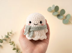 A hand holding a small crocheted plush character that resembles a cute ghost or snowman, wearing a light green scarf, against a warm beige background with soft eucalyptus leaves.