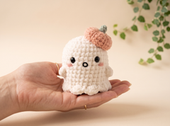 A small crocheted ghost plush toy with a peach pumpkin hat is held in a person's hand against a soft beige background with blurred green leaves.