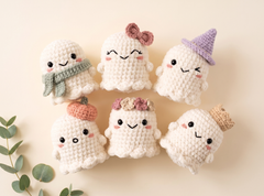 Six cute crocheted ghost plush toys arranged on a warm beige background with a small green leaf sprig at the lower left. The ghosts are white with embroidered faces and wear different accessories including a green scarf, pink bow, purple hat, orange pumpkin, floral crown, and beige crown.
