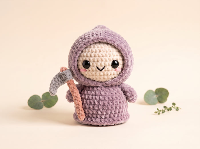 A crocheted amigurumi doll wearing a lavender-purple hooded robe and holding a gray and peach pickaxe, set against a warm beige background with decorative eucalyptus leaves.