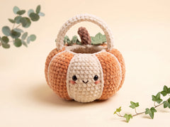 A cute crocheted pumpkin basket with a smiling face on the front, featuring orange and cream stripes, a cream handle, and a small brown stem with green leaves on top, styled on a warm beige background with green ivy and eucalyptus sprigs.