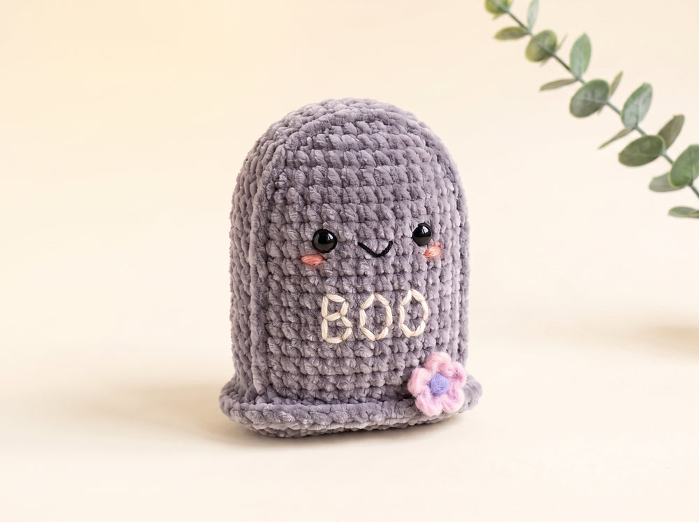 A cute, hand-crocheted ghost-shaped item in lavender-gray with embroidered eyes, a smiling mouth, the word 'BOO' in cream, and a small pink flower accent, set against a warm beige background with a decorative green leaf branch in the corner.