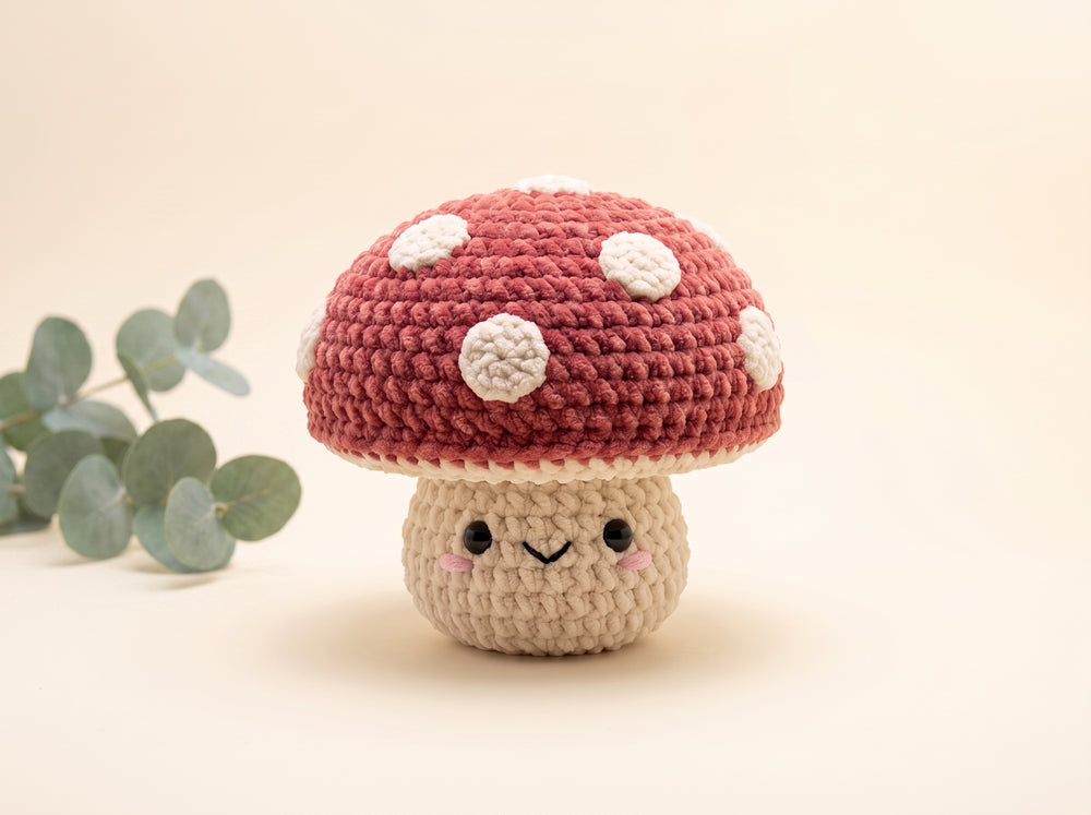 A cute crocheted mushroom toy with a red cap, white spots, and a beige stem with a smiling face, set against a warm beige background with a eucalyptus leaf on the left.