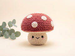 A cute crocheted mushroom toy with a red cap, white spots, and a beige stem with a smiling face, set against a warm beige background with a eucalyptus leaf on the left.
