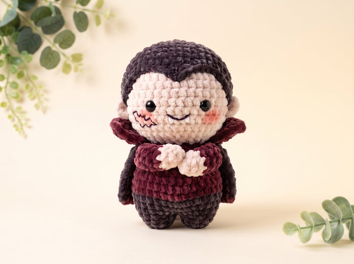 A crocheted vampire doll with dark purple hair and cape, cream-colored face and hands, and a dark burgundy cape, standing against a warm beige background with decorative green leaves.