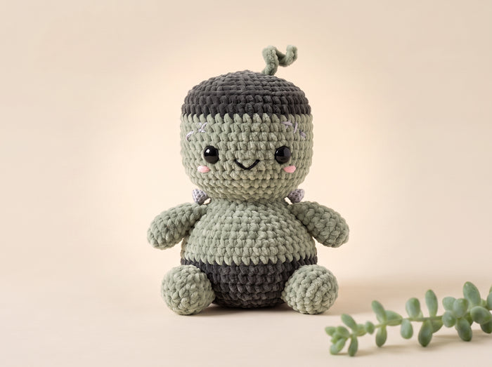 A crocheted amigurumi character doll with a green and dark gray color scheme, styled on a warm beige background with a small green plant sprig in the foreground.