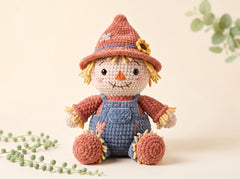 A crocheted scarecrow doll with a brown hat, blue overalls, and yellow hair, posed on a light beige background with soft green plant sprigs in the corners.