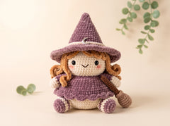 A crocheted witch doll with a purple hat and dress, cream-colored face and legs, and brown curly hair, posed on a warm beige background with soft green eucalyptus leaves.