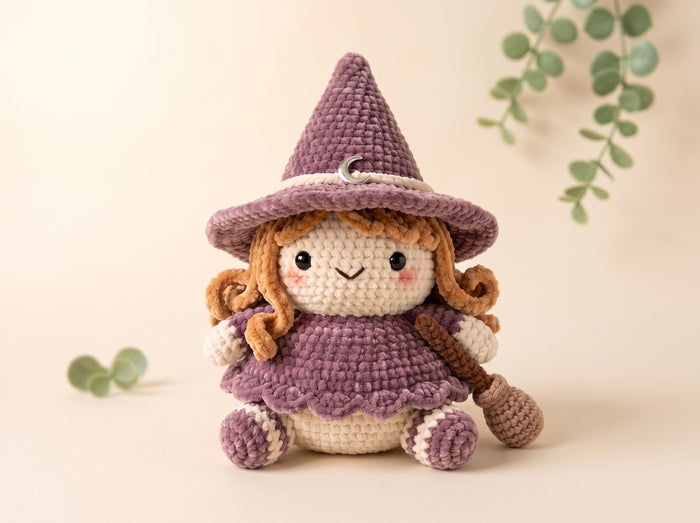 A crocheted witch doll with a purple hat and dress, cream-colored face and legs, and brown curly hair, posed on a warm beige background with soft green eucalyptus leaves.