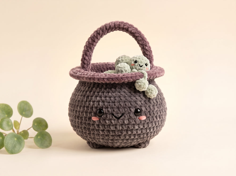 A cute crocheted basket-shaped character toy in dark gray with a purple handle and rim, featuring embroidered facial features and holding a small green plush frog, set against a warm beige background with a decorative green plant in the corner.