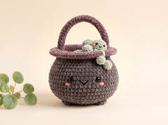 A cute crocheted basket-shaped character toy in dark gray with a purple handle and rim, featuring embroidered facial features and holding a small green plush frog, set against a warm beige background with a decorative green plant in the corner.