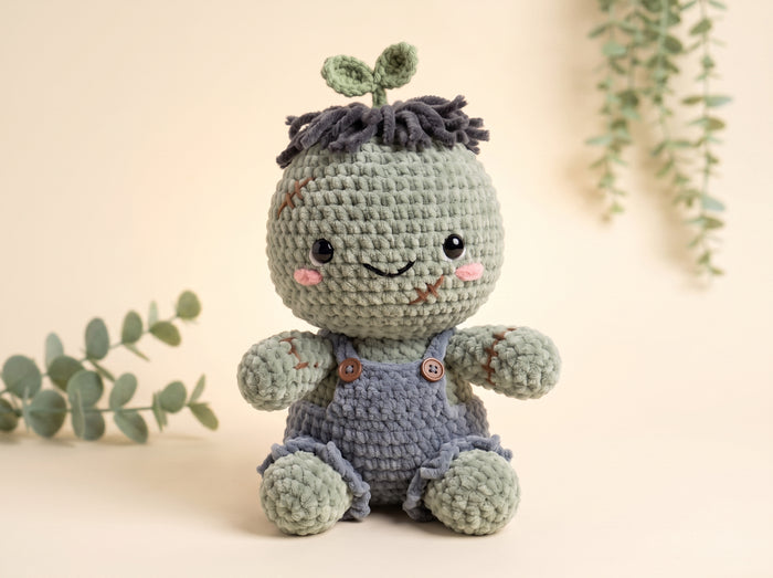 A crocheted plush toy shaped like a cute plant or seedling, sitting on a warm beige surface with soft eucalyptus leaves in the background. The toy has a sage green head with dark gray yarn hair, embroidered facial features, and blue-gray overalls with small brown buttons.