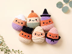 Six cute crocheted candy corn dolls with Halloween-themed hats and bandages, arranged on a light beige background with decorative green leaves in the corners.