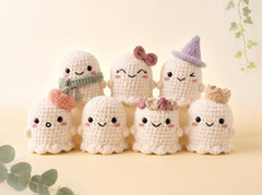 Seven cute crocheted plush ghost toys in off-white, each with different accessories like scarves, hats, bows, and crowns, arranged on a light beige surface with a soft cream background and decorative green leaves.