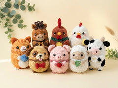 A group of seven crocheted farm and animal plush toys arranged together on a light beige surface with soft decorative leaves in the background. The toys include a brown bear, a brown chicken, a white hen, a pink pig, a white sheep, a black-and-white cow, and an orange cat.