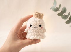 A hand holding a small crocheted plush character with a beige crown, black embroidered eyes and smile, and pink cheeks against a soft cream background with a green leaf branch in the corner.