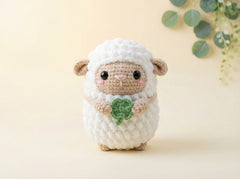 A crocheted plush sheep toy with a white textured body, beige face and ears, pink cheeks, holding a green clover, centered on a warm cream background with soft green leaves in the upper right.