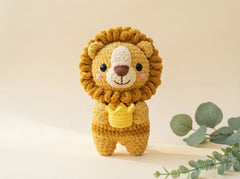 A crocheted lion amigurumi toy holding a small yellow crown, standing against a warm beige background with soft eucalyptus leaves in the corner.