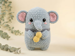 A crocheted gray elephant plush toy holding a yellow peanut, set against a warm beige background with soft eucalyptus leaves in the corners.