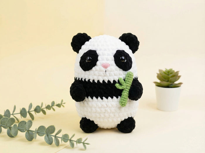 A crocheted panda plush toy holding a green bamboo shoot, set against a warm cream background with decorative eucalyptus leaves and a small potted succulent.