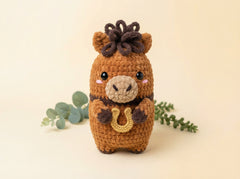 A crocheted toy horse with a brown body, dark brown mane, beige snout, and a small gold horseshoe, photographed on a warm cream background with eucalyptus sprigs.