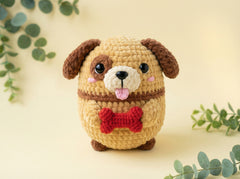 A crocheted plush dog toy with brown floppy ears, a beige body, a red bone-shaped collar, and a pink tongue, centered on a warm cream background with soft green eucalyptus leaves around it.