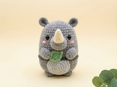 A crocheted gray rhinoceros plush toy holding a green leaf, set against a soft beige background with a small green leaf prop in the corner.