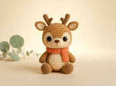 A crocheted plush toy deer with brown and cream yarn, wearing an orange scarf, set against a warm beige background with a small eucalyptus sprig on the left.