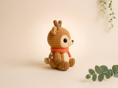 A crocheted stuffed deer toy with brown and cream yarn, wearing an orange scarf, sitting on a warm beige background with decorative green leaves in the corners.