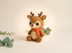 A crocheted plush deer toy with brown antlers and legs, a cream-colored belly and face, and an orange knit scarf, sitting on a warm beige background with decorative eucalyptus sprigs.