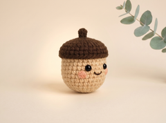 A cute crocheted acorn doll with a smiling face, black eyes, pink cheeks, and a brown cap, set against a warm beige background with a eucalyptus branch in the corner.
