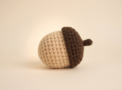 A crocheted acorn toy with a textured beige body and a dark brown cap, shown on a plain beige background.