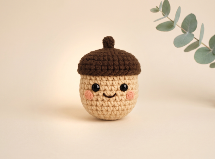 A crocheted acorn character toy with a beige body, brown cap, and embroidered smiling face, centered on a warm cream background with a eucalyptus branch in the upper right.