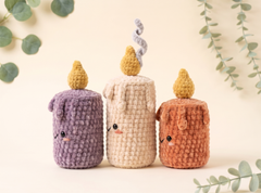Three cute crocheted candle-shaped plush toys in purple, cream, and terracotta orange, arranged on a warm beige background with soft green eucalyptus leaves in the corners.