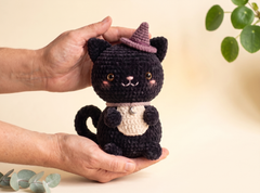A crocheted black cat plush toy with a purple witch hat, cream belly patch, and embroidered facial features, being held gently in two hands against a soft beige background with blurred green leaves.