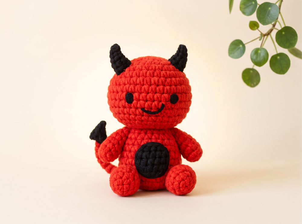 A crocheted devil doll in bright red with black horns, a black tail tip, and a black circular patch on its belly, posed against a warm cream background with a green leafy branch in the upper right.
