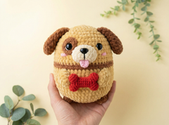 A hand holds a crocheted plush dog toy with brown floppy ears, a cream muzzle, a red knitted bow tie, and a pink tongue, set against a warm beige background with soft green eucalyptus leaves.