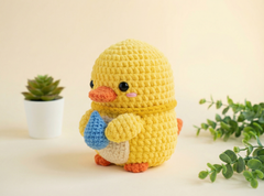 A yellow crocheted duck doll with an orange beak and feet, holding a blue egg, standing on a light beige surface with a small potted succulent and green leaves in the background.