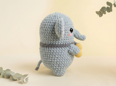 A crocheted plush elephant toy in light blue-gray yarn, holding a small yellow acorn, photographed on a warm cream background with eucalyptus sprigs in the corners.