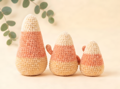 Three crocheted candy corn dolls in different sizes, arranged on a warm beige background with soft green eucalyptus leaves in the upper left corner.