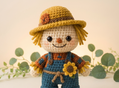 A crocheted scarecrow doll with a yellow hat, blue overalls, and a sunflower detail, set against a warm beige background with soft green leaves.