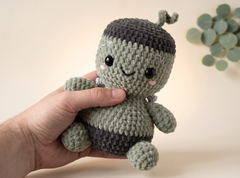 A handmade crocheted amigurumi toy character held in a person's hand against a warm beige background with a soft-focus eucalyptus leaf in the corner.
