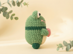 A handmade crocheted plush toy shaped like a cute green frog, standing upright on a soft beige background with decorative eucalyptus leaves in the corners.