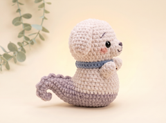 A crocheted plush toy shaped like a small dog or puppy with a tail, shown on a soft cream background with a eucalyptus branch in the corner. The toy is primarily off-white with a lavender tail and a blue collar.
