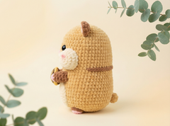 A crocheted hamster toy in warm beige and brown, holding a small brown object, photographed against a soft cream background with eucalyptus leaves around it.