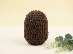 A dark brown crocheted plush toy with small white ears, photographed on a light cream background with a sprig of green eucalyptus leaves in the lower right corner.