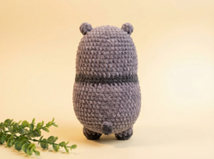 A handmade crocheted plush toy shaped like a small bear or pig, shown from the back on a warm beige background with a green leafy sprig in the lower left corner.