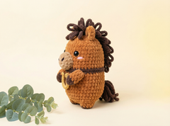 A crocheted plush horse toy in warm brown with a dark brown mane, tail, and ears, holding a small beige and brown accessory. The toy is photographed on a soft cream background with a sprig of green leaves at the lower left.