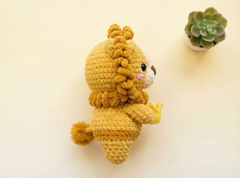 A crocheted lion toy in mustard yellow and cream, shown from the back on a warm cream background with a small potted succulent in the upper right.